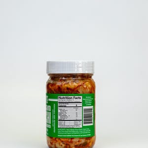 May include: A glass jar of kimchi with a white lid and a green label. The label displays nutrition information and ingredients. The kimchi is a mix of red and white vegetables. The jar is labelled "PERISHABLE KEEP REFRIGERATED".