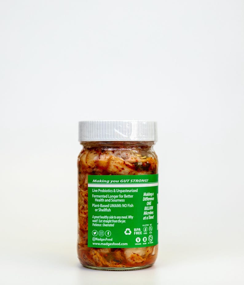 May include: A glass jar with a white lid, filled with a reddish-orange fermented food product. The jar has a green label with text including "Making you GUT STRONG!" and "Live Probiotics & Unpasteurized". The product is plant-based and contains no fish or shellfish.