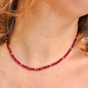 Op de afbeelding: Een delicate ketting met kleine, gefacetteerde, dieprode kralen afgewisseld met kleine goudkleurige kralen. De ketting wordt om de hals gedragen en toont zijn eenvoudige elegantie en levendige kleur.