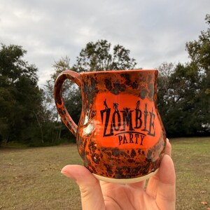 May include: Orange ceramic mug with black speckled design and a handle. The mug features the words "ZOMBIE PARTY" in black lettering. The mug is held in a hand against a background of green grass and trees.