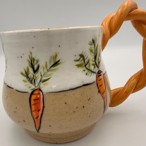 May include: A ceramic mug with a white upper section and a speckled tan lower section, featuring hand-painted orange carrots with green leafy tops. The mug includes a distinctive, thick orange handle shaped like a twisted rope or braid.