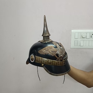 May include: Black and bronze military helmet with a tall, pointed spike on top. The helmet features ornate bronze detailing, including an eagle emblem and decorative bands. A historical piece.