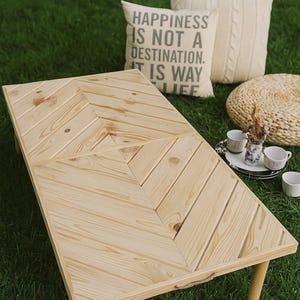 Può includere: Un tavolino da caffè in legno chiaro con un motivo a spina di pesce su un prato verde. Il tavolo ha quattro gambe corte. Sono visibili anche due cuscini decorativi e un piccolo vassoio con tazze. Un cuscino ha il testo "HAPPINESS IS NOT A DESTINATION. IT IS WAY LIFE."