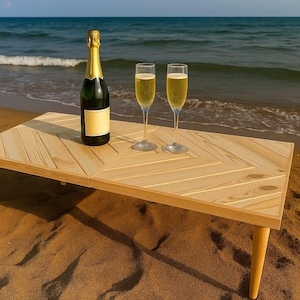 May include: A light wood picnic table on a sandy beach, with a bottle of champagne and two glasses. The table is made of light-colored wood and the sea is visible in the background. The table is approximately 90 cm long.