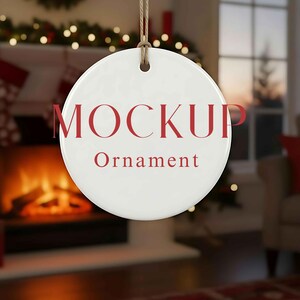 Könnte beinhalten: Ein weißer, runder Ornament mit dem Wort "MOCKUP" in roter Schrift über dem Wort "Ornament". Das Ornament hängt an einer braunen Schnur. Der Hintergrund zeigt einen Kamin und Weihnachtsdekorationen.