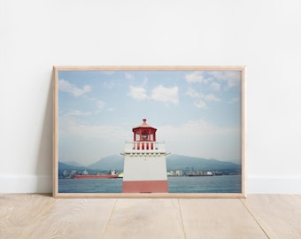 May include: Framed print of a lighthouse with a red top and base, set against a blue sky with clouds. The lighthouse is white with red accents, situated near a body of water with mountains in the background. A ship is visible in the distance.