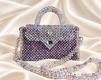 May include: A structured handbag crafted from iridescent, multi-faceted beads in shades of purple and clear crystal. The bag features a top handle, a fold-over flap with a silver-toned metal logo plate, and a matching beaded shoulder strap.