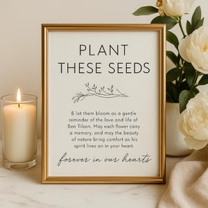 May include: A framed print with gold trim, featuring the text "PLANT THESE SEEDS" and a floral illustration. The text below reads "forever in our hearts". A lit candle and a vase of white flowers are in the background.