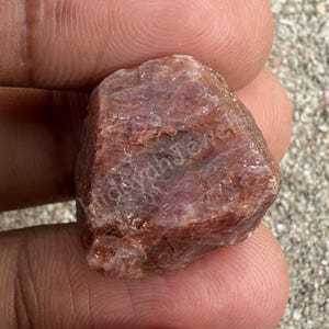 Puede incluir: Una gema de rubí en bruto, sin cortar, de color marrón rojizo. La superficie de la piedra tiene un aspecto texturizado, con líneas e imperfecciones visibles. La imagen muestra la forma natural y en bruto del rubí, destacando su forma orgánica y variaciones de color. El texto "Hidayah Jewel" es visible.
