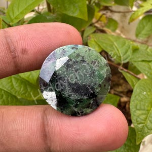 Puede incluir: Una gema redonda y facetada con una mezcla de tonos verde oscuro y negro. La gema se sostiene entre dos dedos, con un fondo de hojas verdes. La superficie de la gema refleja la luz, mostrando sus facetas pulidas.
