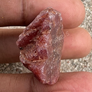 May include: A natural, rough-cut ruby gemstone in a deep red shade. The raw mineral has a textured surface and an irregular shape, demonstrating its natural, unpolished form. The ruby is held in a hand, emphasizing its size and colour.