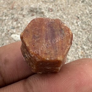 May include: A raw, reddish-brown gemstone with a faceted surface. The stone is held in a hand, with the text "Hidayah Jewels" visible on the side. The gemstone appears to be a rough, uncut mineral.