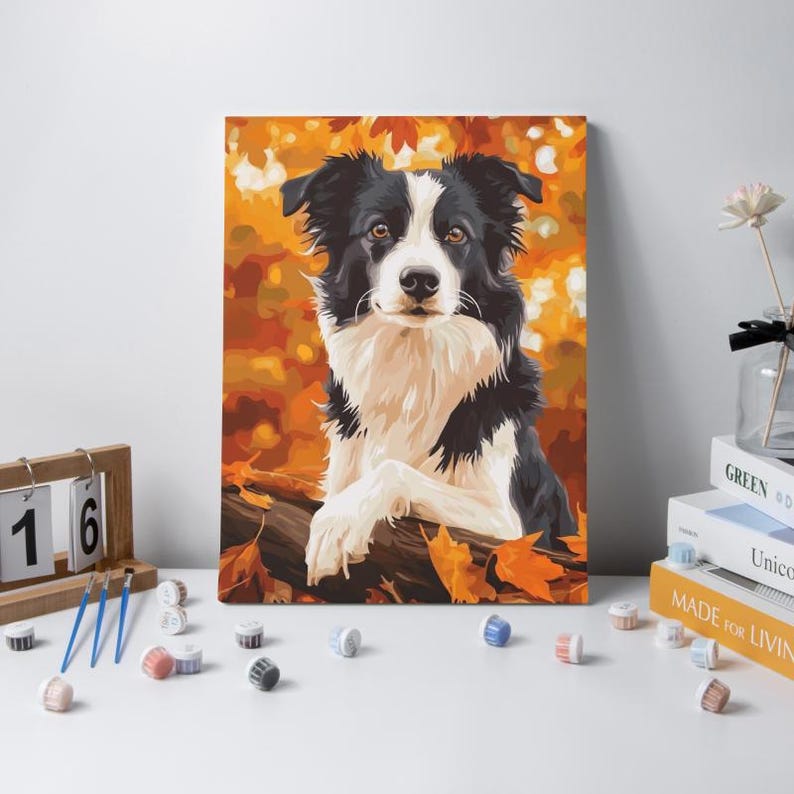 May include: A paint-by-numbers canvas featuring a black and white Border Collie resting on a log amidst a vibrant autumn forest background with orange and yellow leaves. The artwork is displayed alongside paintbrushes and small paint pots.