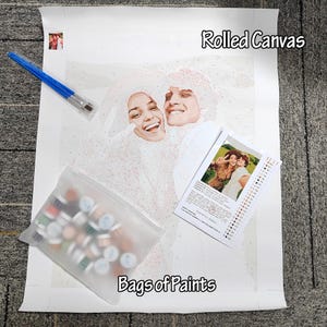 May include: A paint-by-numbers kit featuring a rolled canvas with a smiling couple, a set of paintbrushes, a bag of paints, and a reference photo. The canvas is labeled "Rolled Canvas" and the bag is labeled "Bags of Paints".