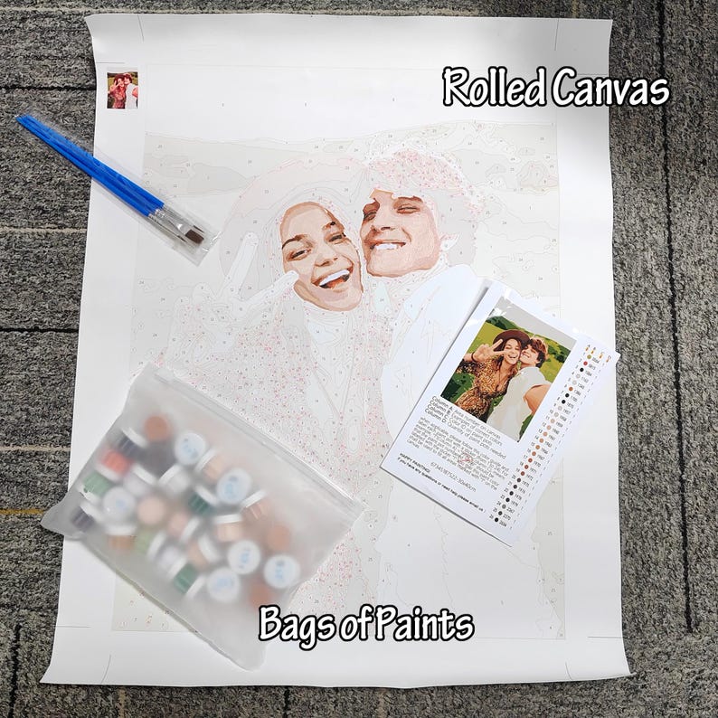 May include: A paint-by-numbers kit featuring a rolled canvas with a smiling couple, a set of paintbrushes, a bag of paints, and a reference photo. The canvas is labeled "Rolled Canvas" and the bag is labeled "Bags of Paints".