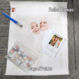 May include: A paint-by-numbers kit featuring a rolled canvas with a smiling couple, a set of paintbrushes, a bag of paints, and a reference photo. The canvas is labeled "Rolled Canvas" and the bag is labeled "Bags of Paints".