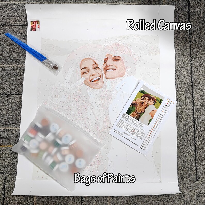 May include: A paint-by-numbers kit featuring a rolled canvas with a smiling couple, a set of paintbrushes, a bag of paints, and a reference photo. The canvas is labeled "Rolled Canvas" and the bag is labeled "Bags of Paints".