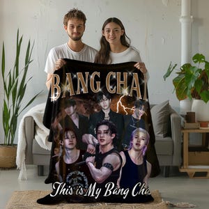 May include: Black blanket with the gold text "BANG CHAN" and images of a musical group. The blanket also has the text "This is My Bang Chan" in white. A man and a woman are holding the blanket. The blanket is a decorative item.