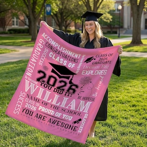 May include: A pink graduation blanket with black and white text. The blanket says "Class of 2026", "William", and "You Did It!" with graduation cap graphics. The blanket is held by a person wearing a graduation gown.
