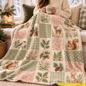 Könnte beinhalten: Eine weiche Wohndecke mit einem Patchwork-Design aus abwechselnden Quadraten in Salbeigrün, Creme und zartem Rosa. Das Muster zeigt Illustrationen von Waldtieren wie einem Reh, einem Eichhörnchen und einem Hasen sowie Tannenzapfen, Eicheln und Blumen.
