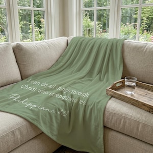 Può includere: Una coperta verde salvia drappeggiata su un divano. La coperta presenta un testo bianco che recita: "Tutto posso in colui che mi fortifica. Filippesi 4:13."