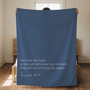May include: A navy blue blanket featuring a quote from Isaiah 40:31 in white text. The quote reads, "But those who hope in the Lord will renew their strength. They will soar on wings like eagles." The blanket is displayed indoors.