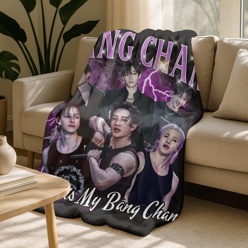 May include: A black and purple throw blanket featuring a collage of images of a musical group. The blanket has the text "Bang Chan" and "is My Bang Chan" in white and purple. The blanket is draped over a beige sofa.