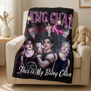May include: Black blanket with a group of six people and the text "ANG CHAN" in purple and white. The blanket also has the text "This is My Bang Chan" in white. The blanket is draped over a beige armchair.