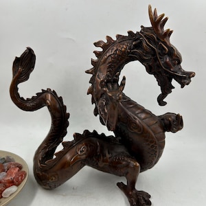 May include: A detailed bronze dragon sculpture with intricate scales, horns, and a flowing mane. The dragon is posed in a dynamic, curved position, with its tail curled and head raised. The sculpture is a rich, dark brown color.