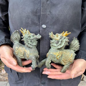 May include: Two antique bronze-colored mythical creatures, possibly Qilin, are held in hands. The figures have detailed features, including horns, manes, and ornate detailing on their bodies. The creatures are a mix of green and gold.