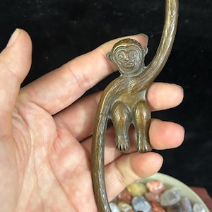 May include: A bronze-colored monkey figurine with a curved tail, designed to hang. The monkey is depicted in a seated position, with its arms extended to hold onto the tail. The figurine has detailed facial features and a textured surface.