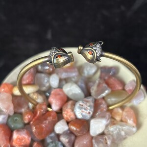 May include: A gold-toned bracelet with two fox head terminals. The fox heads have orange eyes and are embellished with small, clear crystals. The bracelet is displayed on a bed of colorful, polished stones, with a dark background.