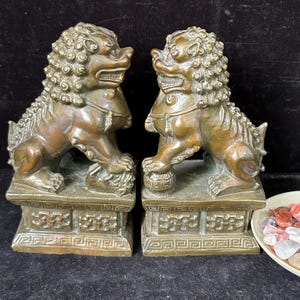 May include: A pair of bronze-colored guardian lion statues, each mounted on a rectangular pedestal with geometric carvings. One lion rests a paw on a small cub, while the other rests a paw on a spherical ball. Both figures feature curly manes.