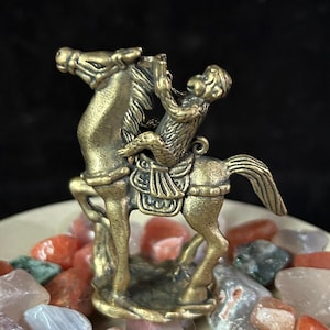 May include: A bronze-colored figurine of a monkey riding a horse. The monkey is holding a small object, and the horse has detailed saddle and bridle. The figurine is set on a bed of colorful stones, with a dark background.