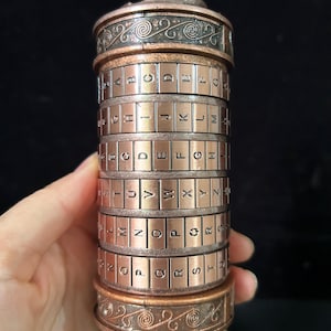 May include: A cylindrical copper-toned cryptex puzzle with rotating rings featuring embossed letters of the alphabet. The top and bottom bands display intricate swirling patterns, while the central sections contain rows of letters for code setting.