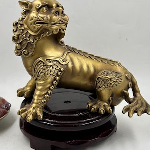 May include: A brass-colored mythical creature figurine with a lion-like head, scaled legs, and a long tail, standing on a dark brown circular wooden base. The creature features intricate relief details on its mane, limbs, and back, with a metallic finish.