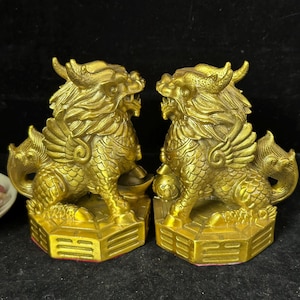 May include: A pair of gold-colored statues depicting mythical creatures with wings, scales, and horns. Each creature stands on a hexagonal base featuring engraved lines, with one creature resting a paw on a round object and the other on a gold ingot.