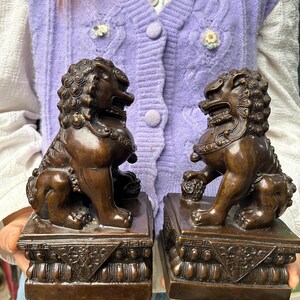 May include: A pair of bronze-colored guardian lion statues, each seated on a rectangular base with intricate relief carvings. The lions feature detailed manes, open mouths, and muscular forms, with one lion resting a paw on a spherical object.