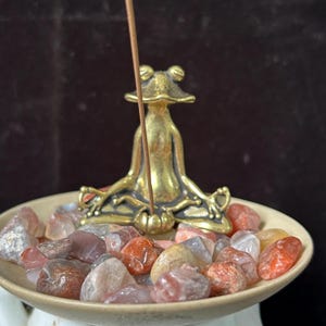 May include: A brass frog incense holder in a seated yoga pose. The frog sits on a beige ceramic dish filled with small, colorful tumbled stones. An incense stick is inserted into the holder. The background is a dark brown.