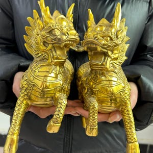 May include: Two golden, mythical creature statues with detailed scales, horns, and open mouths. The statues are held in front of a dark background, showcasing their intricate design and metallic sheen. The figures are in a seated position.