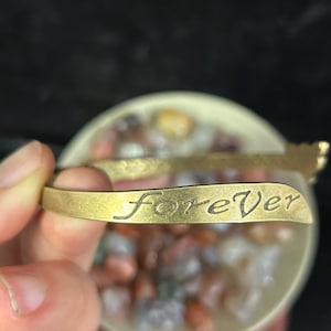 May include: A gold-colored metal bracelet with the word "forever" engraved on the front. The bracelet is curved and has a simple design. The background is blurred, with a bowl of colorful stones.