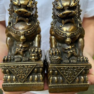 May include: A pair of bronze-colored guardian lion statues, each featuring intricate facial details, a collar with a pendant, and a textured base with decorative carvings. One lion rests a paw on a small cub, while the other rests a paw on a sphere.