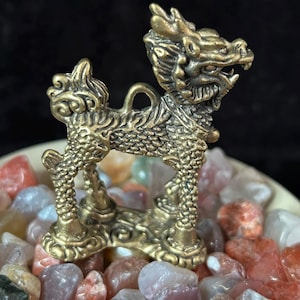 May include: A detailed brass figurine of a mythical dragon-like creature. The sculpture is standing on a bed of colorful polished stones. The dragon has intricate details, including scales and a fierce expression. The figurine is approximately 3 inches tall.