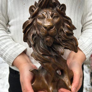 May include: Bronze lion sculpture with a detailed mane and facial features. The sculpture is held in front of a white sweater. The lion is mounted on a textured base, showcasing a regal and powerful aesthetic.