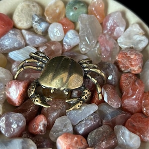 May include: A detailed brass crab figurine rests amidst a collection of polished gemstones in shades of red, orange, gray, and clear. The crab has intricate detailing on its shell and legs, creating a striking contrast against the colorful stones.