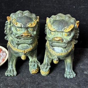 May include: A pair of dark bronze-toned guardian lion statues featuring intricate swirling manes, wide eyes, and bared teeth. Each figure is adorned with gold-colored accents on the forehead, ears, and collar, with one lion resting a paw on a sphere.