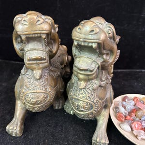 May include: A pair of bronze-colored guardian lion statues with open mouths and detailed chest plates featuring embossed characters. The statues are positioned next to a small, shallow bowl filled with various small, polished, multi-colored stones.