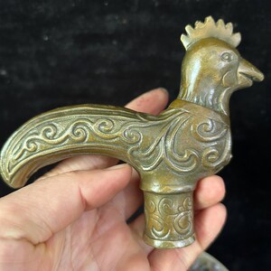 May include: A bronze-colored, decorative rooster figurine. The rooster has detailed feather and wing patterns, a comb, and a beak. The figurine is held in a hand, with a dark background.
