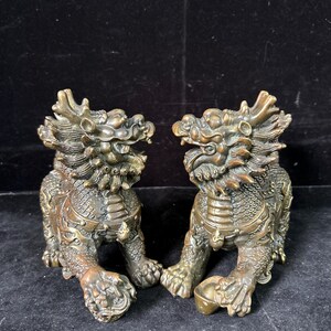 May include: A pair of bronze-colored metal statues depicting mythical creatures with dragon-like heads, scaled bodies, and flowing manes. Each figure is in a crouching stance with intricate detailing on the fur and scales, one resting a paw on a small orb.