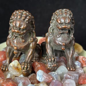 May include: Two bronze-colored Foo Dog statues, each with detailed features, including textured fur and a regal posture. The statues are displayed on a bed of colorful polished stones, creating a decorative display.
